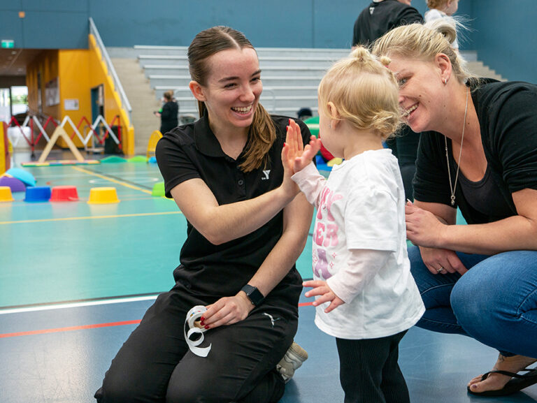 A smiling Y WA staff member gives a high five to a toddler while the child's parent looks on warmly during an indoor sports session