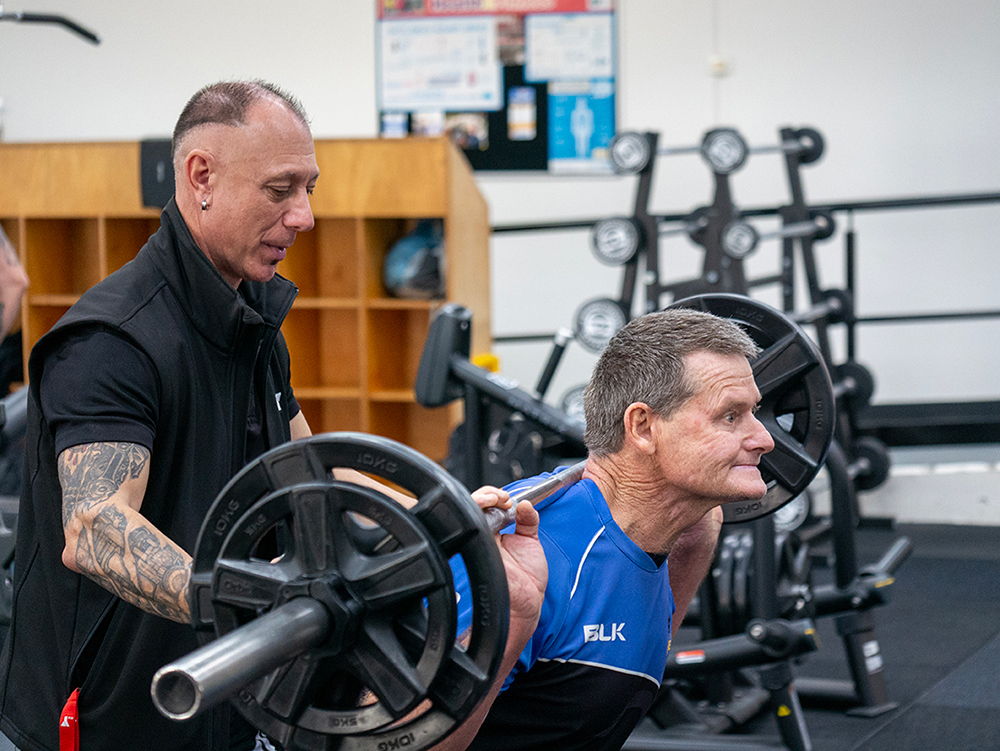A personal trainer spots an older man performing a barbell squat in a gym weight room