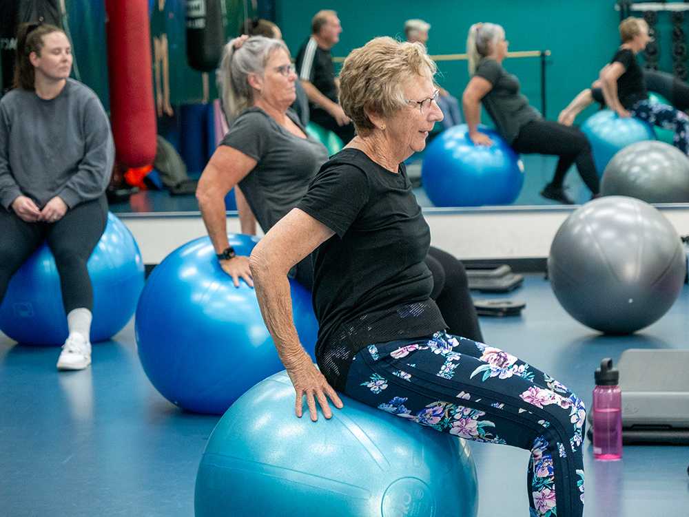 A group of women of varying ages sit on exercise balls during a group fitness class in a gym studio