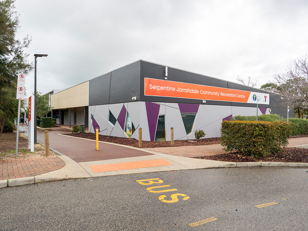 Exterior of the Serpentine Jarrahdale Community Recreation Centre with a bold orange sign, purple geometric facade detailing, and a bus zone in the foreground
