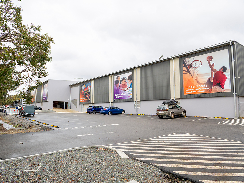 Exterior of a large recreation centre building with large promotional banners advertising indoor sports and outside school hours care, with a car park in the foreground