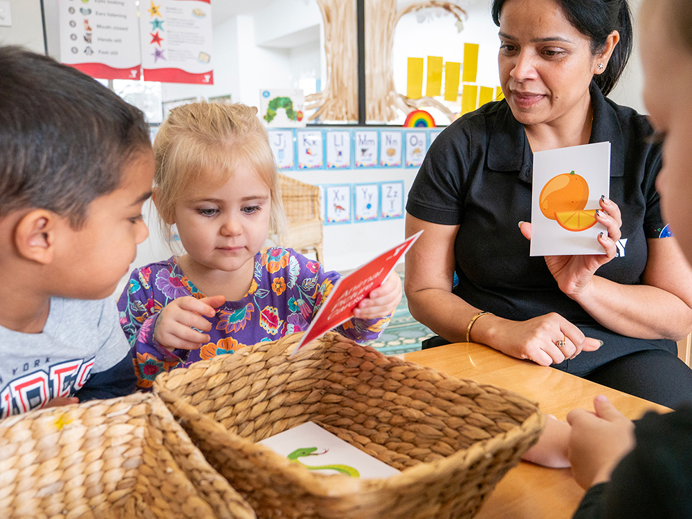 An educator holds up a flashcard of an orange while two young children look at picture cards from a wicker basket in a colourful early learning classroom