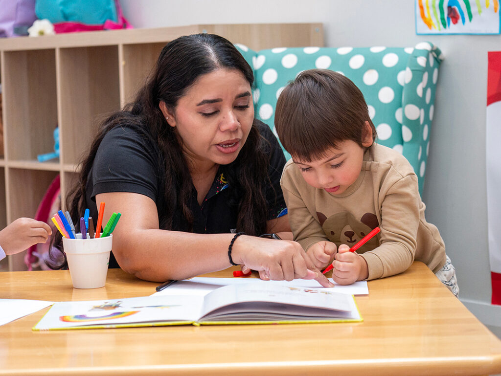 A Y WA educator guides a young child through a drawing activity in a book at a classroom table.