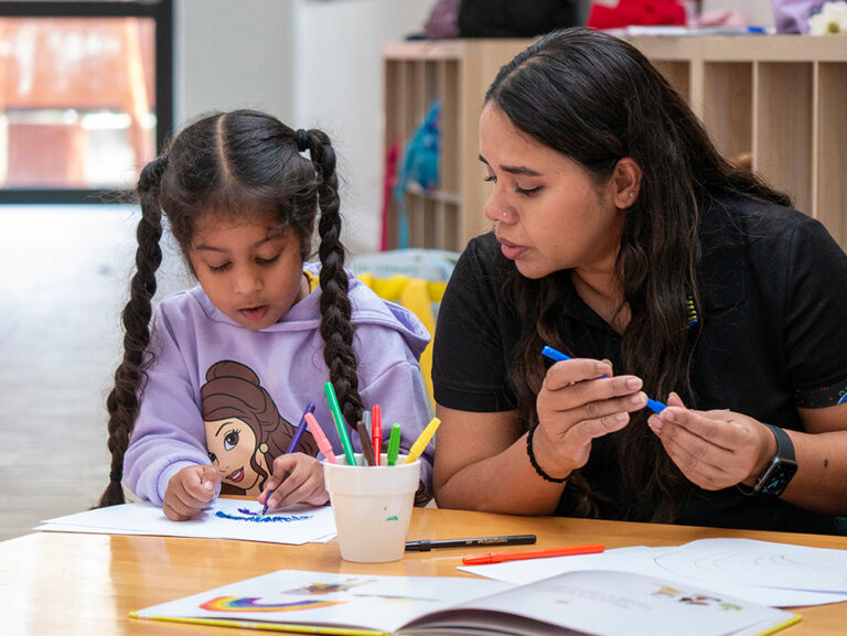 A Y WA educator sits with a young child drawing together at a table with colourful markers.