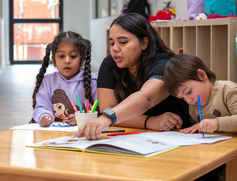 Two children and an educator sitting at a desk reading a book and completing an activity together