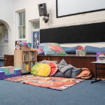 A cosy reading nook with cushions, bean bags, a bookshelf filled with children's books, and an Indigenous-design rug on the floor