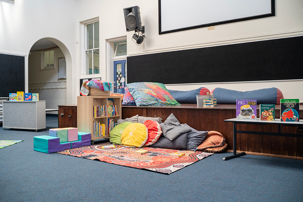 A cosy reading nook with cushions, bean bags, a bookshelf filled with children's books, and an Indigenous-design rug on the floor