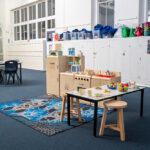 A children's dramatic play kitchen area with wooden toy appliances, food, and crockery set out on a table, sitting on an Indigenous-design rug