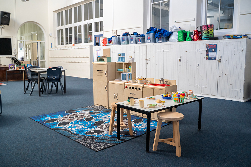 A children's dramatic play kitchen area with wooden toy appliances, food, and crockery set out on a table, sitting on an Indigenous-design rug
