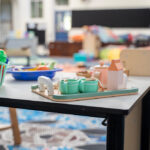 A wooden toy tea set with cups and teapot on a tray, alongside a basket of colourful toy fruit and vegetables on a table