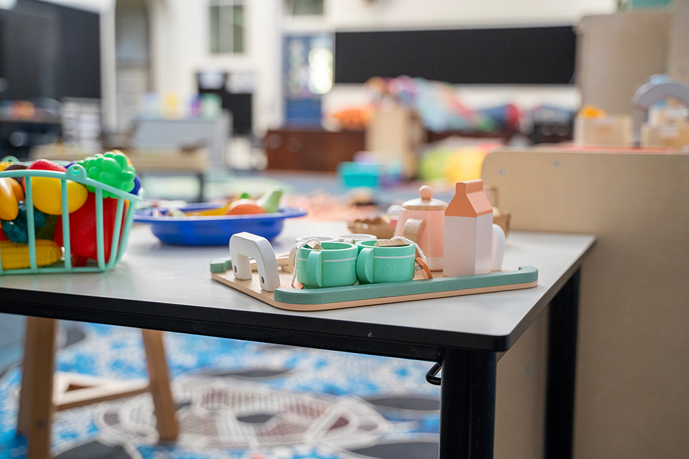 A wooden toy tea set with cups and teapot on a tray, alongside a basket of colourful toy fruit and vegetables on a table