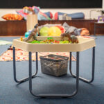 A sensory play table filled with toy dinosaurs and green grass on a stand, set up in a children's activity room