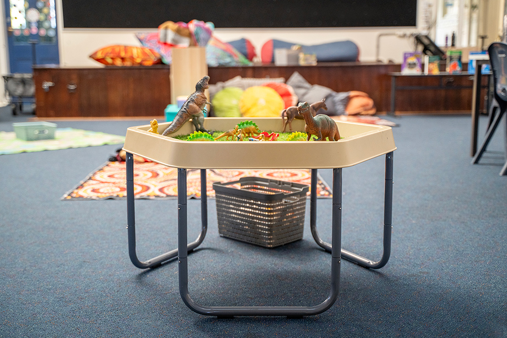 A sensory play table filled with toy dinosaurs and green grass on a stand, set up in a children's activity room