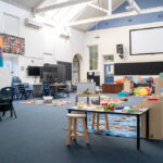 A wide view of a spacious after-school care room with high ceilings, featuring various activity stations including a play kitchen, building toys, reading nook, and stacking cushions