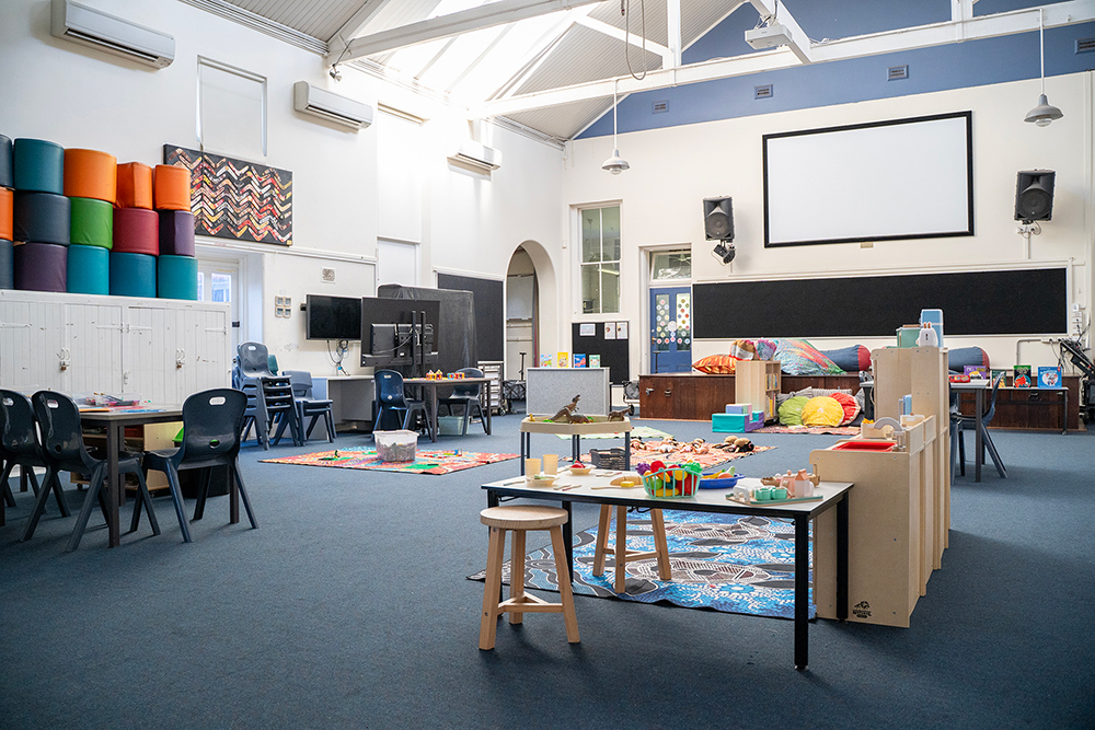 A wide view of a spacious after-school care room with high ceilings, featuring various activity stations including a play kitchen, building toys, reading nook, and stacking cushions