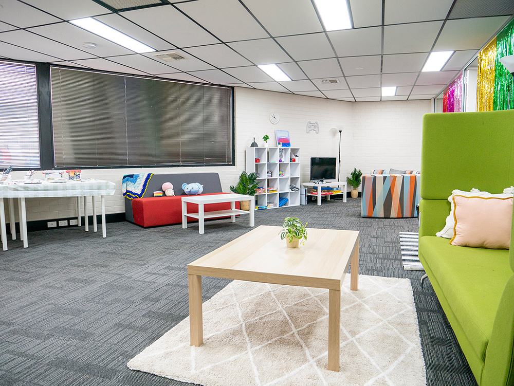 A bright youth drop-in centre lounge room with colourful furniture including a red sofa, green high-back couch, and multicoloured armchair, shelving with board games and toys, a small TV, indoor plants, and a light timber coffee table on a white rug