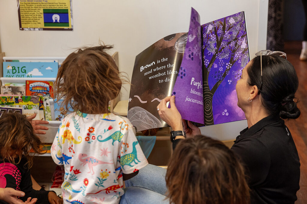 A woman reads a colourful picture book about Australian nature to a group of young children in a playroom