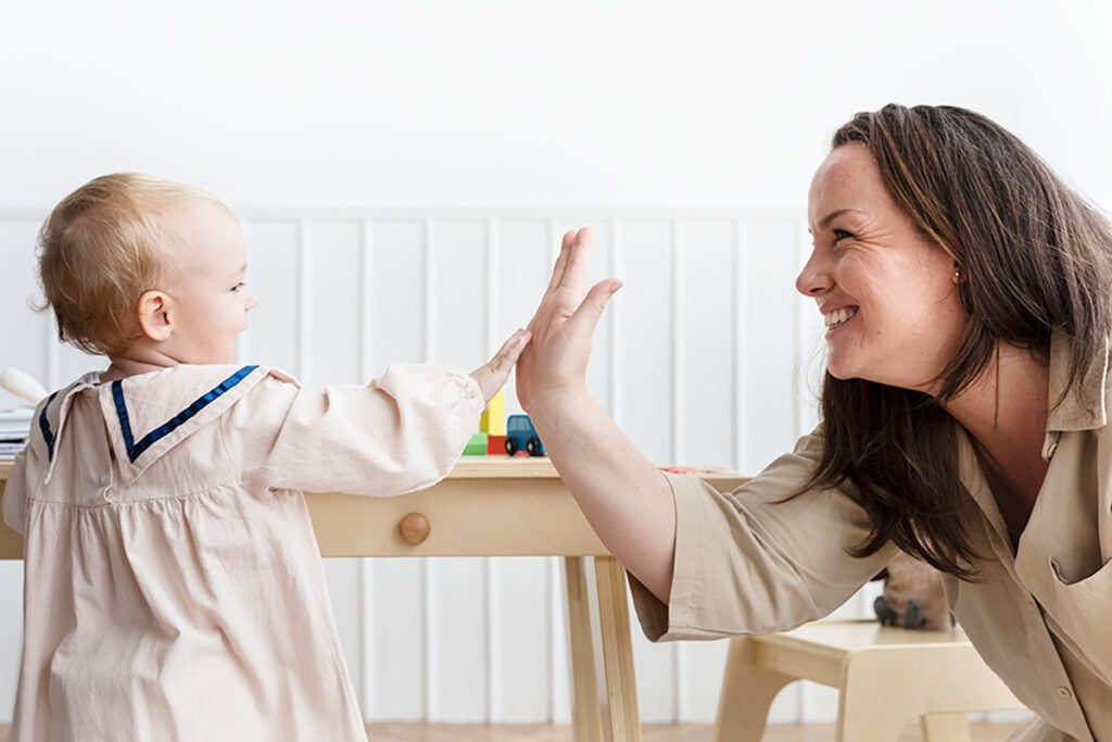 A smiling woman gives a high five to a toddler across a small wooden table in a bright playroom