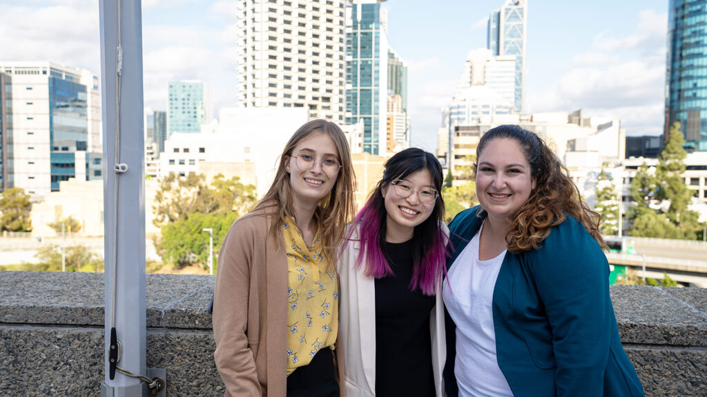 Three young women smile together on a rooftop with the Perth city skyline behind them