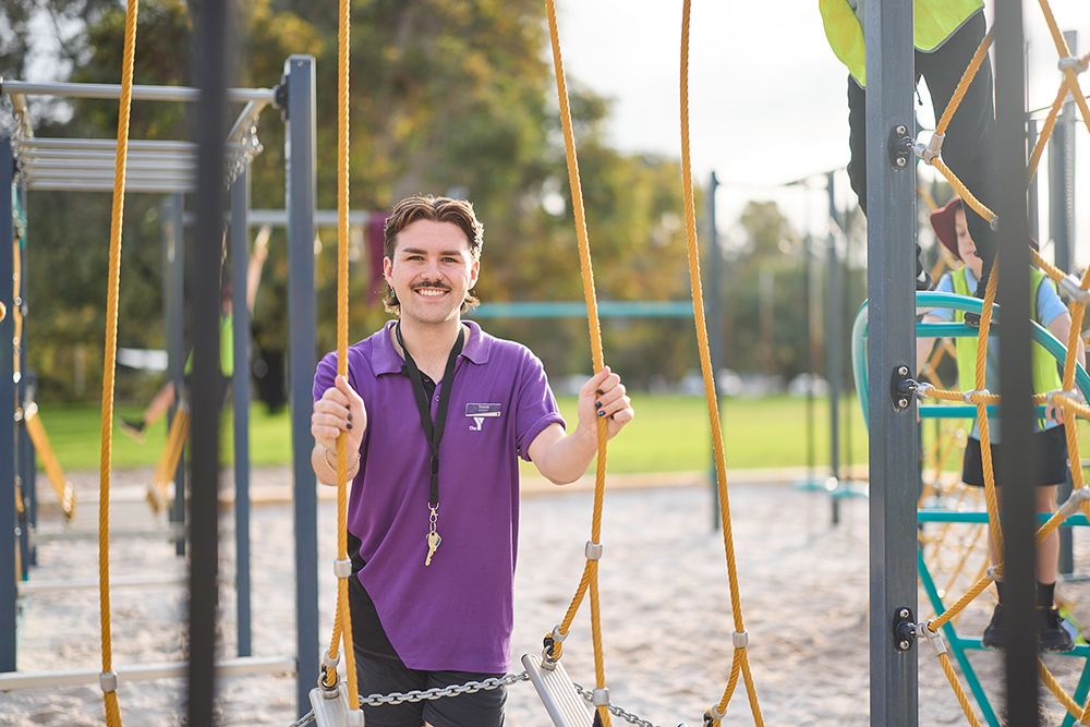 A smiling Y WA staff member in a purple polo shirt stands among the ropes of an outdoor playground climbing structure