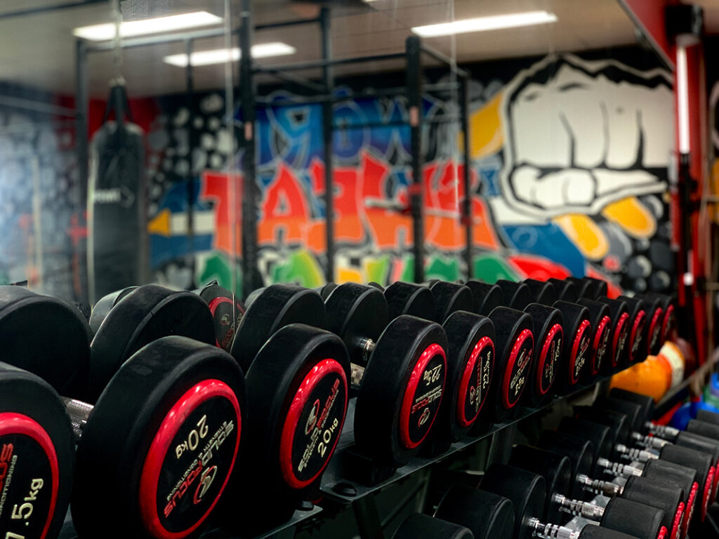 Rows of black and red dumbbells on a rack in a gym, with a colourful graffiti mural and a punching bag visible in the background
