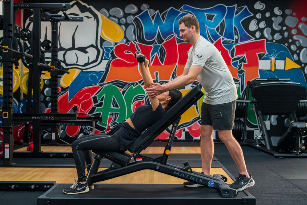 16:19 A personal trainer assists a woman performing a dumbbell exercise on an incline bench, with a bold graffiti mural reading "Work Sweat" on the gym wall behind them
