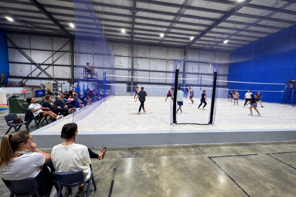 Players compete on an indoor sand volleyball court inside a large warehouse facility, watched by spectators seated courtside