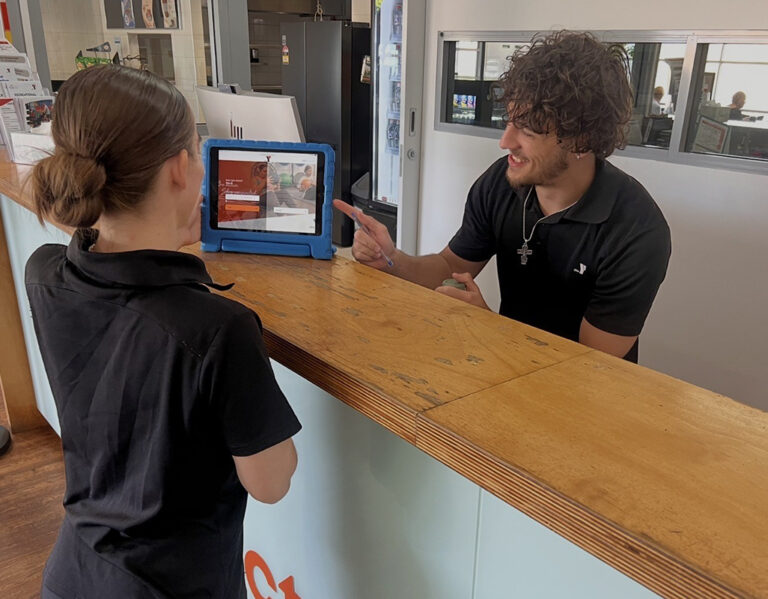 A Y WA staff member smiles as he assists a customer at a reception desk, with a tablet displaying a membership sign-up screen propped between them