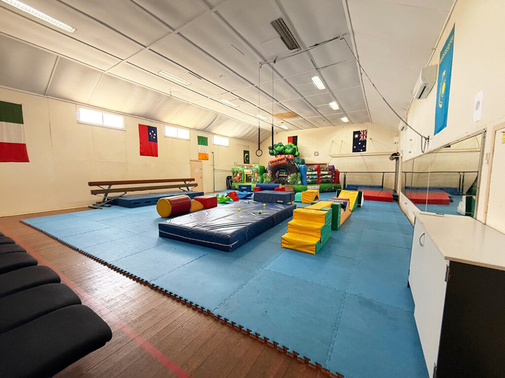 A children's soft play room with foam mats, climbing blocks, a balance beam, and an inflatable play structure, decorated with international flags