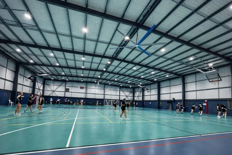 An empty indoor sports stadium with a multi-use court, basketball hoops, tiered spectator seating, and overhead lighting