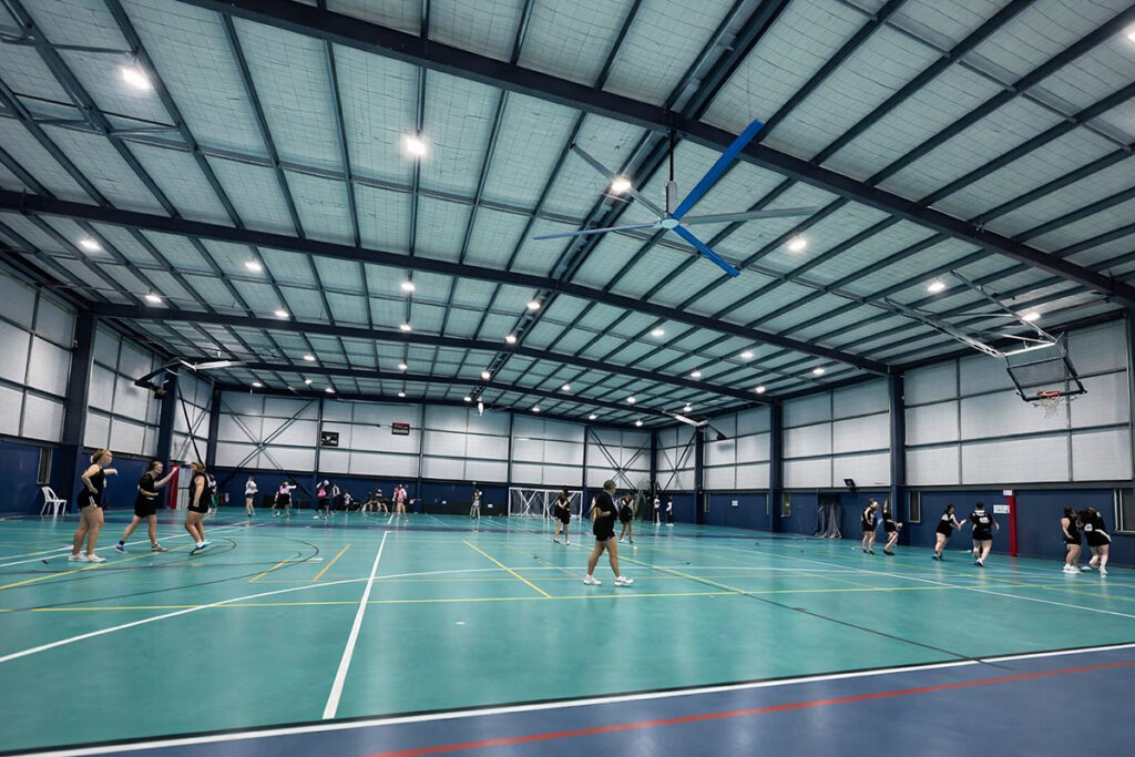An empty indoor sports stadium with a multi-use court, basketball hoops, tiered spectator seating, and overhead lighting