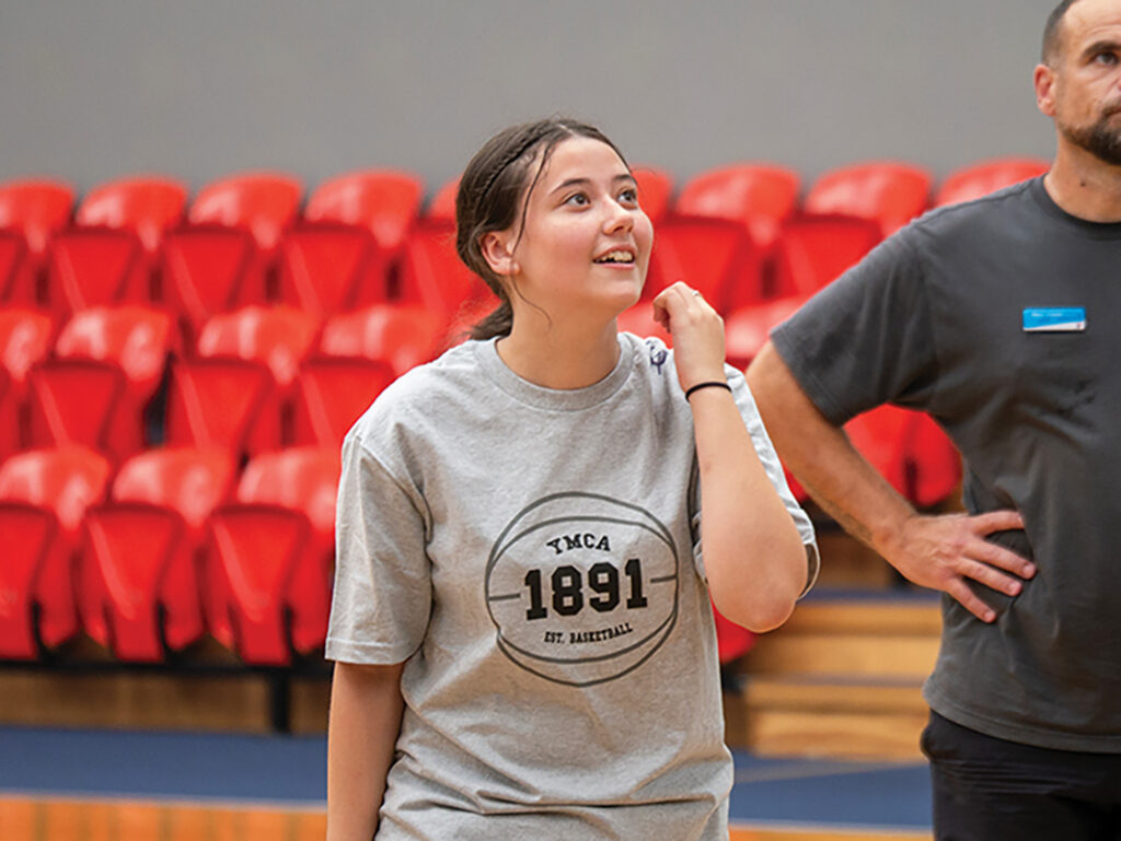 A young female student wearing a YMCA 1891 basketball t-shirt looks up during a basketball session in a school gymnasium