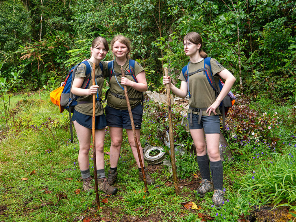 16:42 Three young women in matching khaki shirts and backpacks pause on the Kokoda Trail, holding walking sticks and standing beside remnants of a crashed aircraft in the junglE