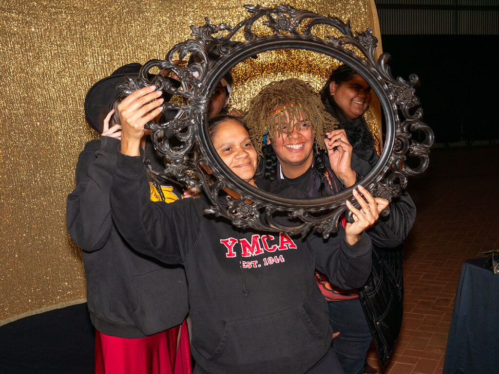 Two people smile and pose together through a decorative oval frame at a NAIDOC Ball photo booth, with a gold sequin backdrop behind them