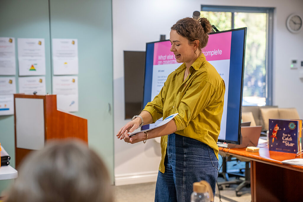 A facilitator in a yellow shirt presents to a group during a School Ready training session, with a slide visible on screen behind her