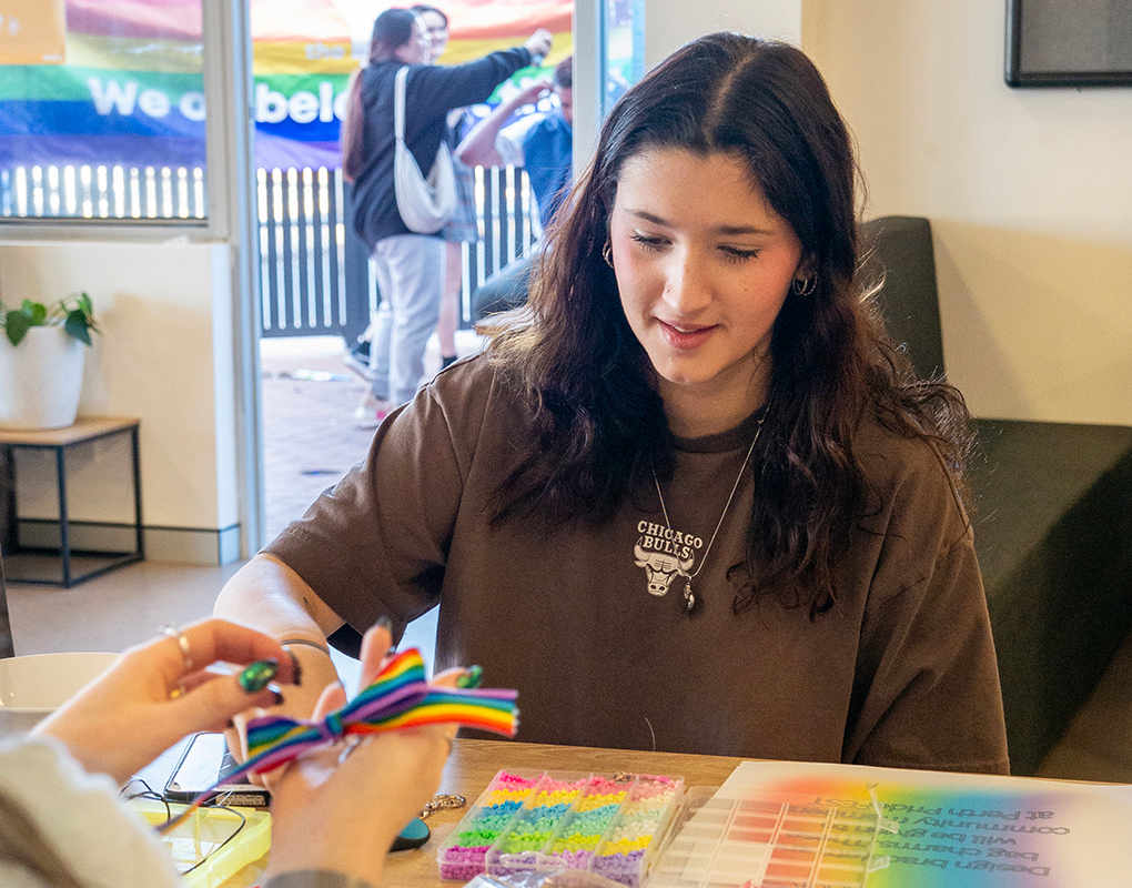 A young person smiles as they receive a rainbow ribbon at a craft table during an IDAHOBIT community fair, with a pride flag and attendees visible through the window behind them