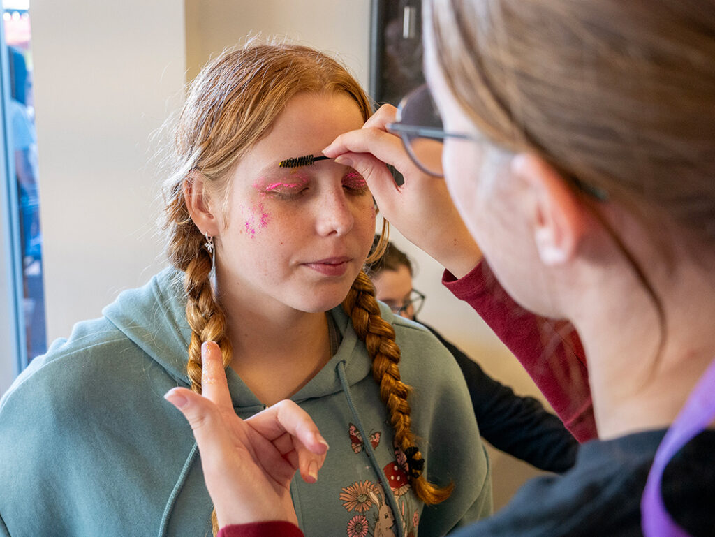 A student with pink and glitter eye makeup has their eyebrows styled by another student at the Y School Mirrabooka IDAHOBIT Fair