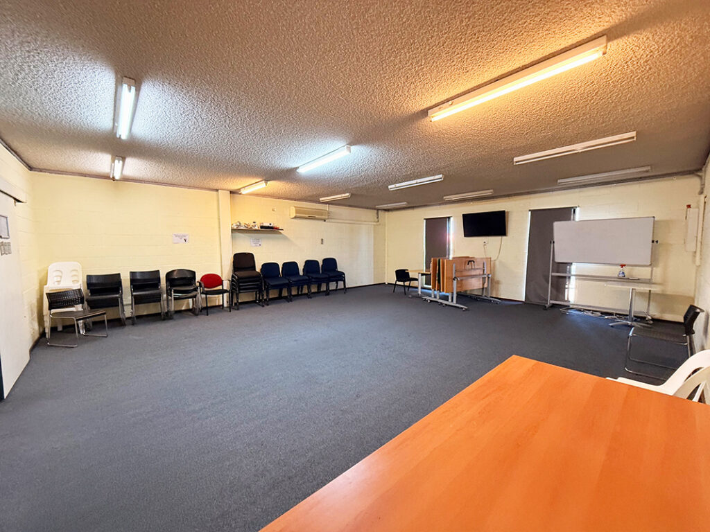 A multipurpose meeting room with rows of chairs along the wall, a wall-mounted TV, whiteboard, and a timber table in the foreground