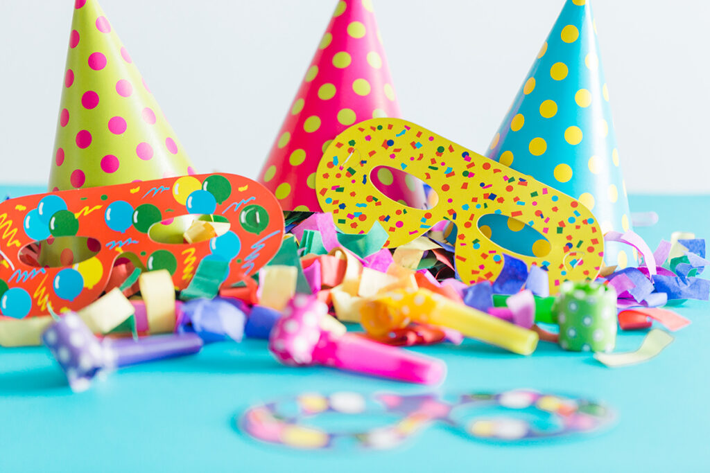 Colourful polka-dot party hats, decorative masks, party blowers, and confetti scattered on a light blue surface