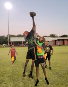 two young people jumping up for a football on a green oval outside