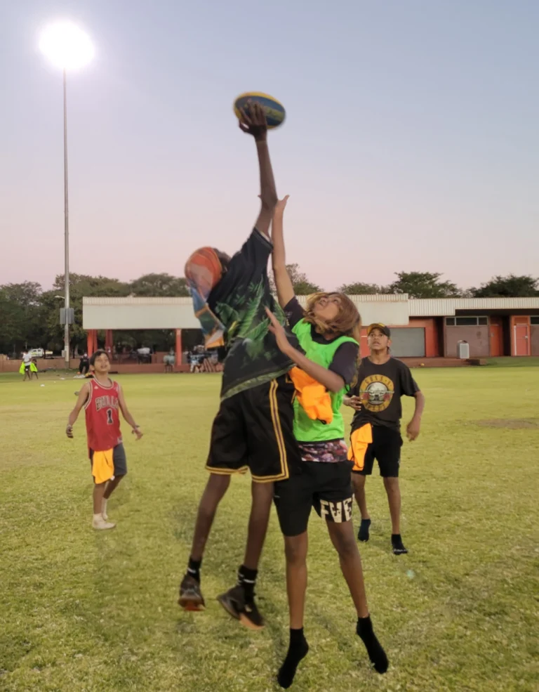 two young people jumping up for a football on a green oval outside