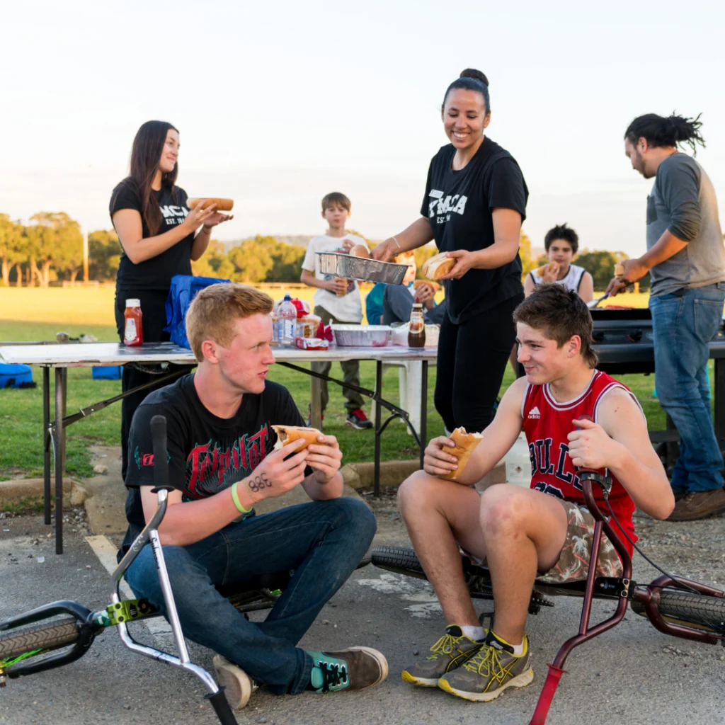 group of young people sitting around a table outside eating hot dogs, laughing and enjoying eachothers company