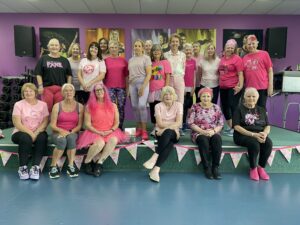 Group of around 20 women in pink outfits and pink accessories smile for a group photo in a fitness studio, with pink bunting along the stage