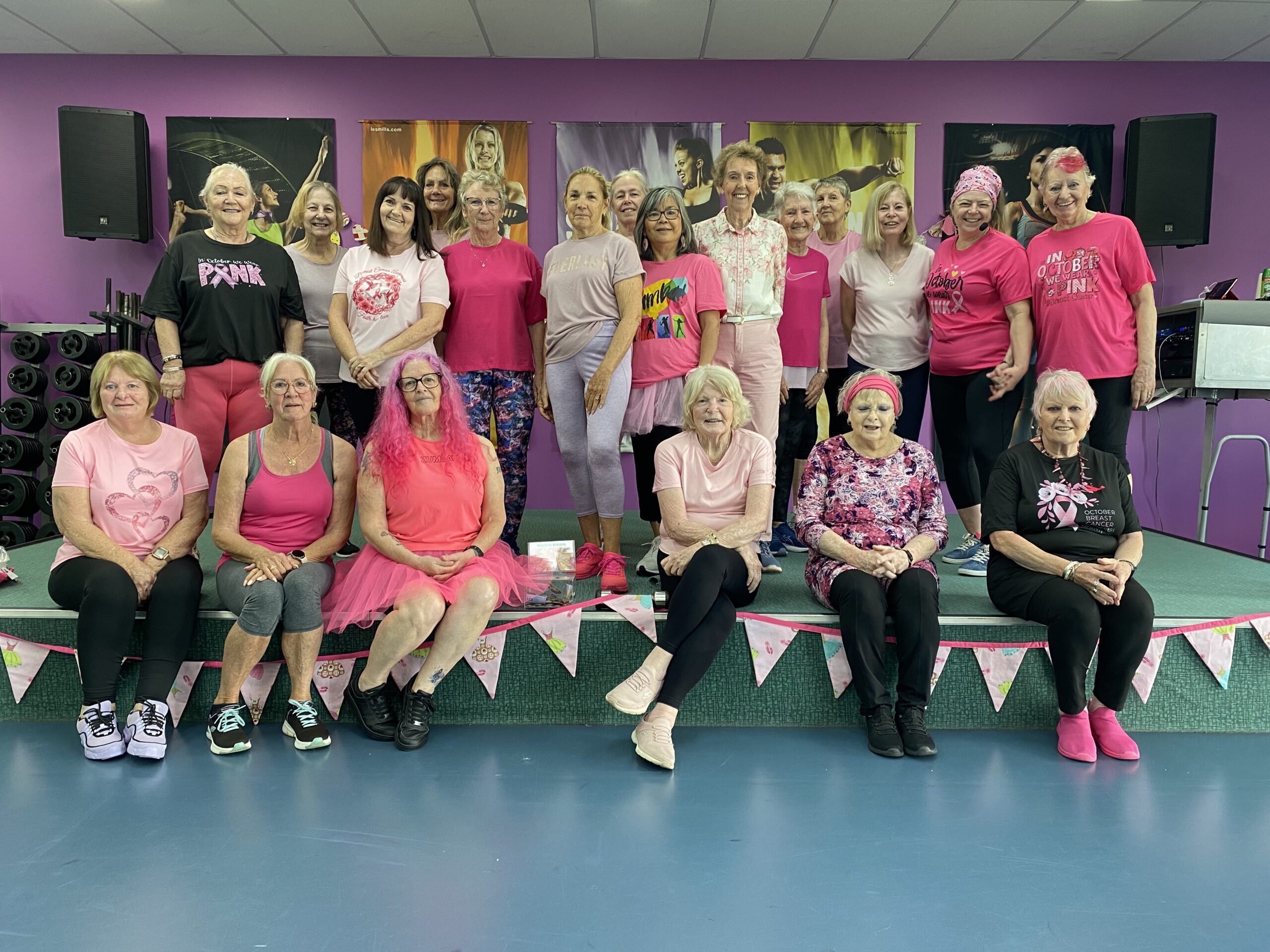 Group of around 20 women in pink outfits and pink accessories smile for a group photo in a fitness studio, with pink bunting along the stage