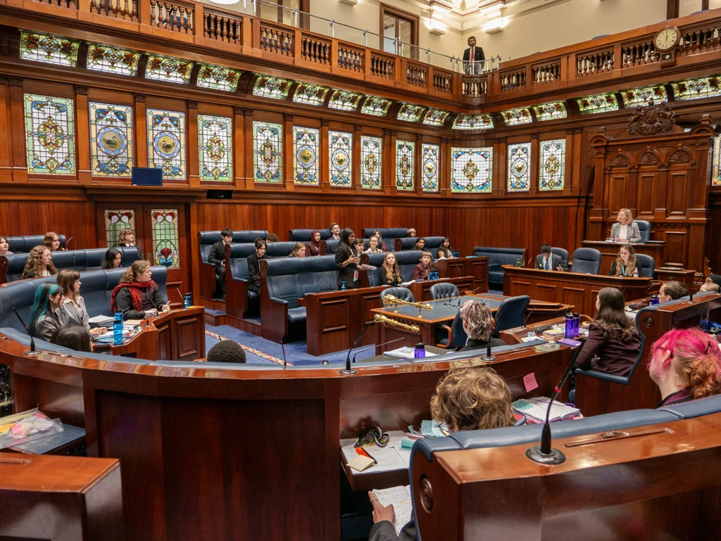 A wide view of a heritage parliamentary chamber with dark timber panelling, stained-glass windows and blue leather seating. A young person speaks from the benches while others listen, with the presiding officer seated at the raised chair on the right.