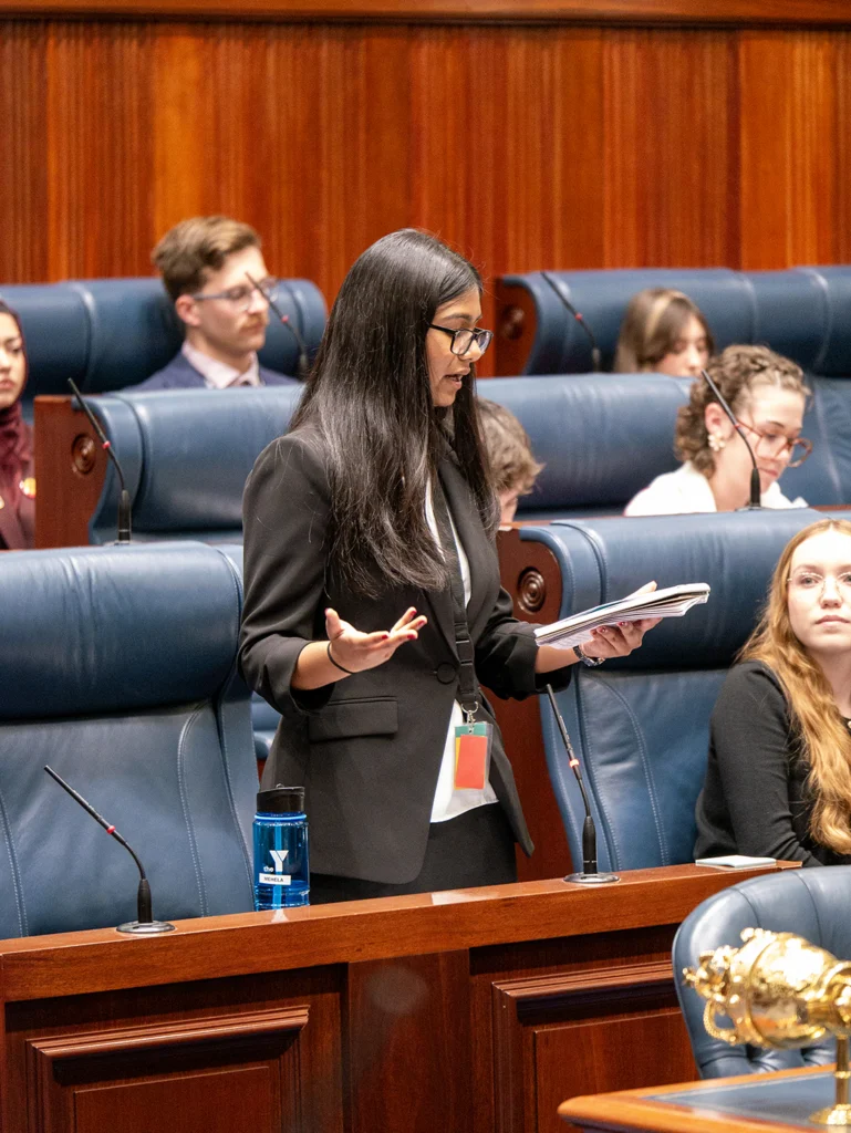 A young woman in a black blazer and glasses stands to speak in a parliamentary chamber, holding papers and gesturing with one hand. Rows of blue leather seats and other seated participants are behind her.