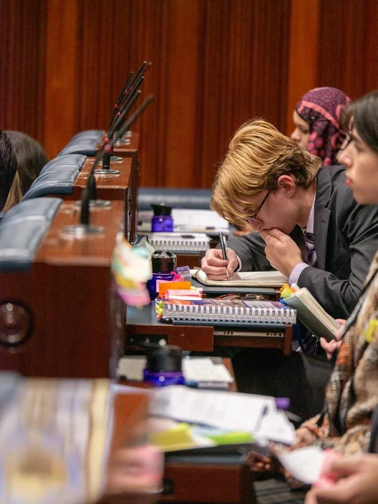 A young person with glasses leans over a desk, writing in a notebook, surrounded by papers, sticky notes and water bottles. Other participants are seated beside and behind them in the chamber.