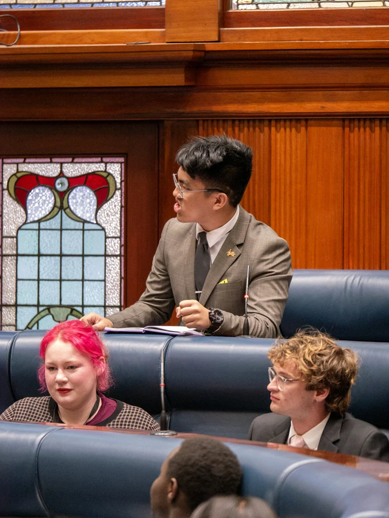 A young man in a grey-brown suit stands at a desk in a wood-panelled chamber, speaking and looking to his left, with a stained-glass panel behind him. Two seated young people in the foreground look on.