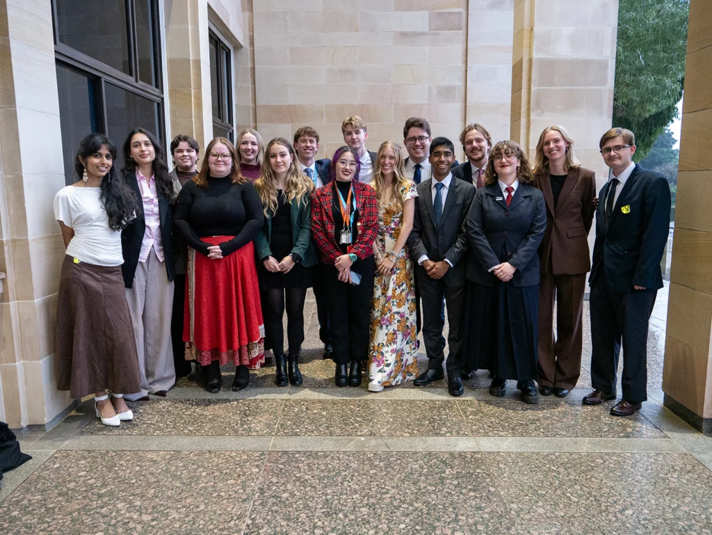 A group of 17 young people in smart and semi-formal clothing stand close together for a group photo under a sandstone archway, smiling at the camera.